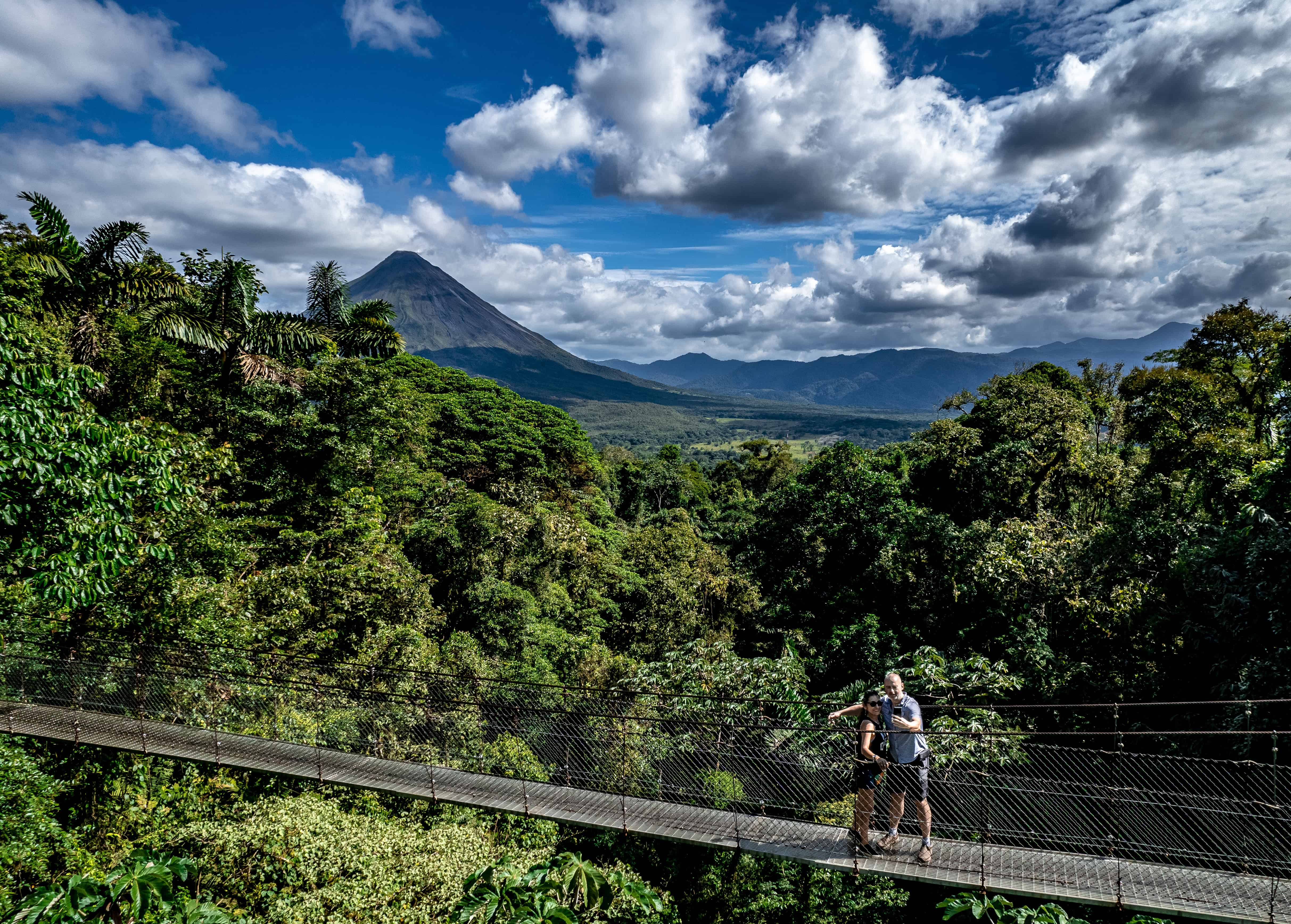 Pareja tomándose una selfie en un puente colgante con el volcán Arenal al fondo.