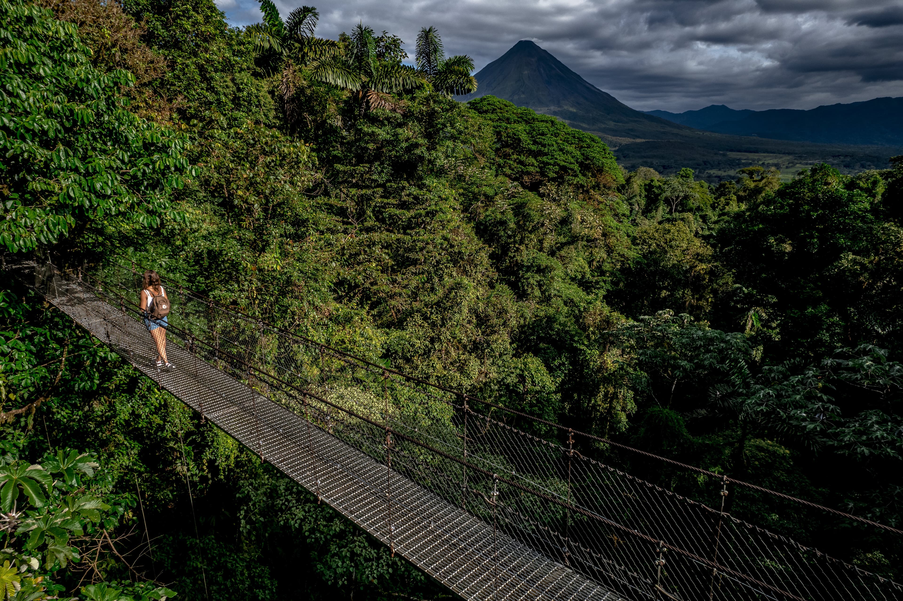 Mujer cruzando un puente colgante sobre la selva tropical con un gran volcán al fondo.