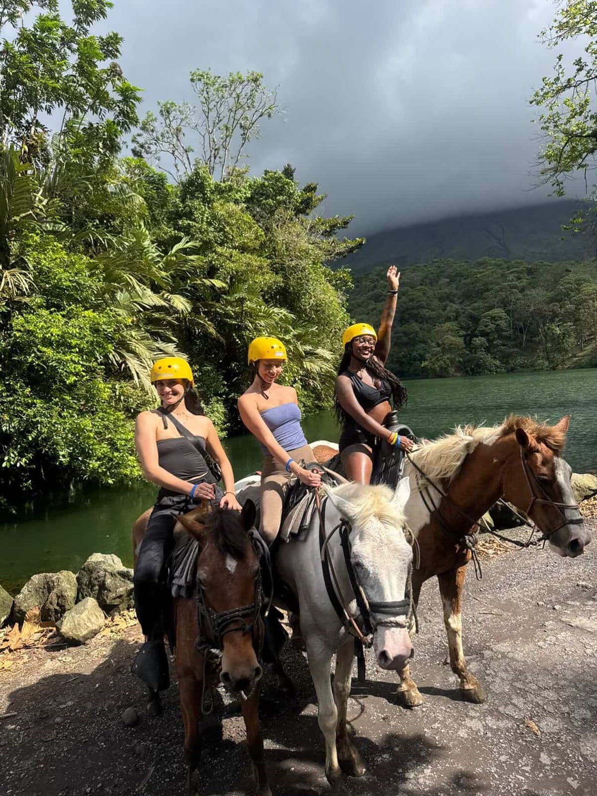Tres mujeres con cascos amarillos montan a caballo frente a un lago y montañas verdes.