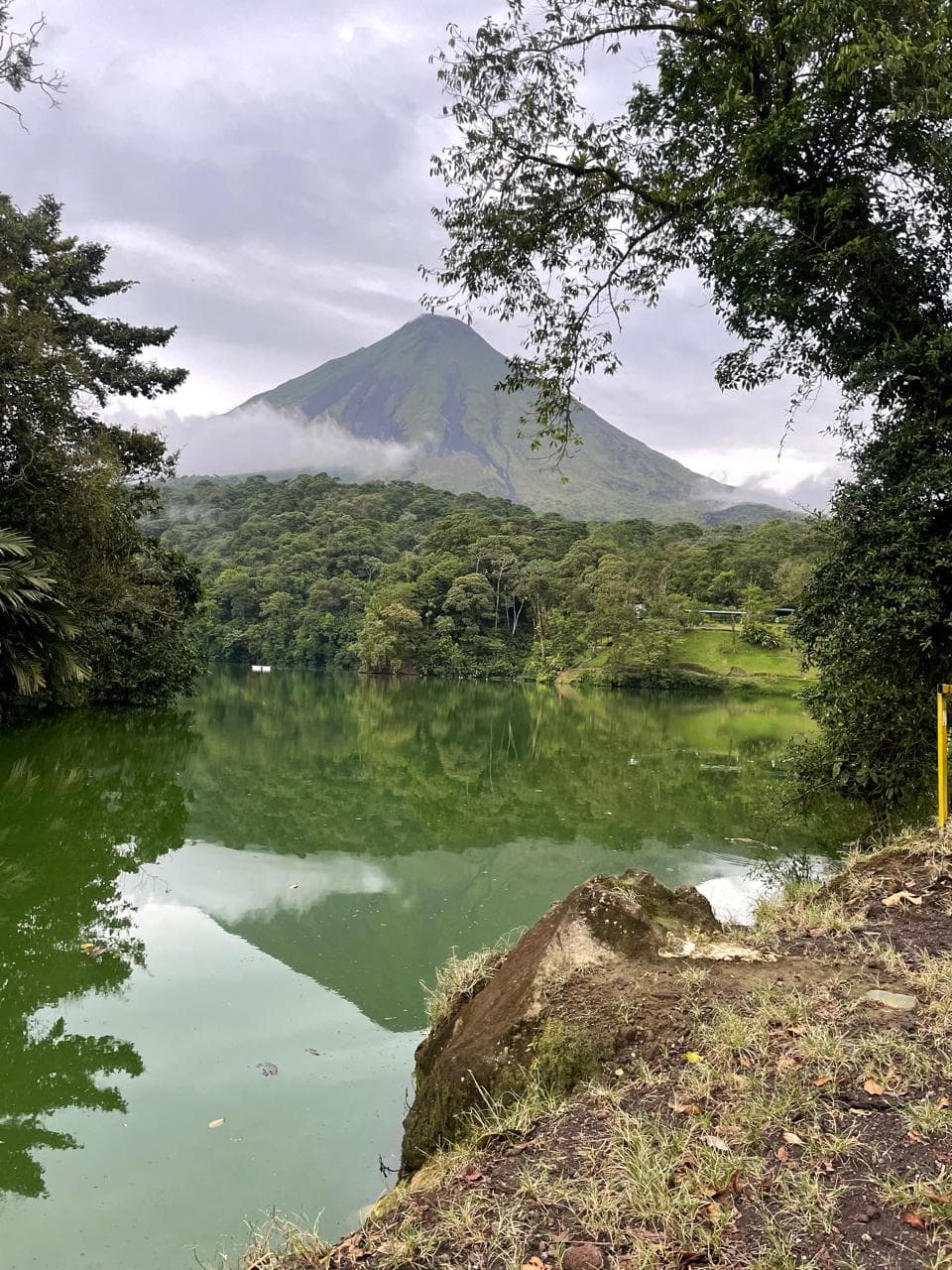 Volcán cónico reflejado en un lago verde rodeado de densa vegetación tropical y nubes.