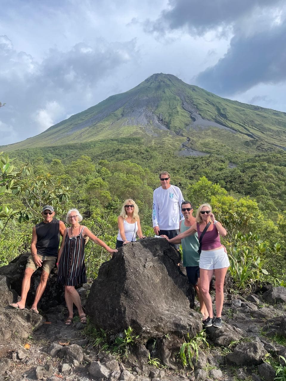 Seis personas posan frente a un gran volcán verde rodeado de exuberante vegetación tropical.