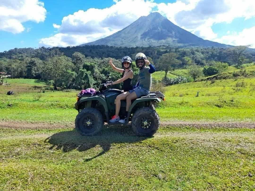 Dos personas en cuatrimoto posan con pulgares arriba frente a un majestuoso volcán cónico.