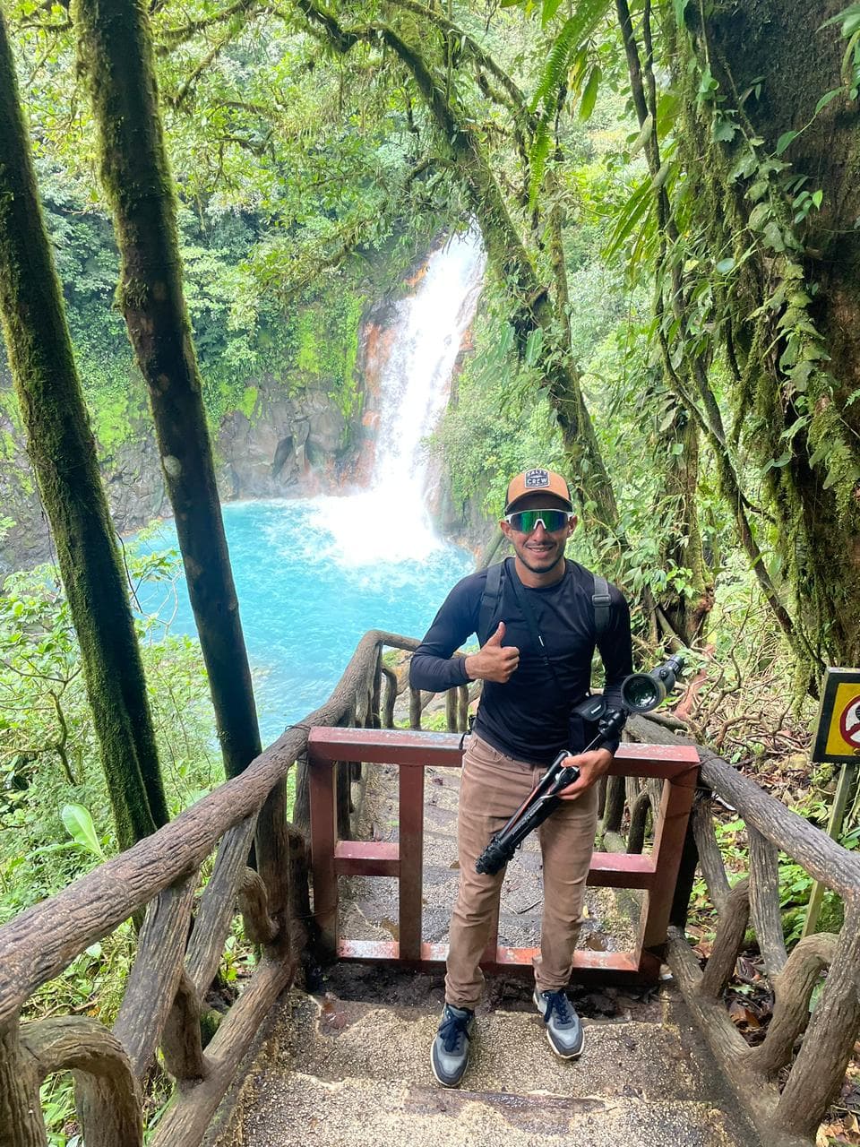 Hombre con equipo de cámara posa frente a una vibrante cascada turquesa en la selva.
