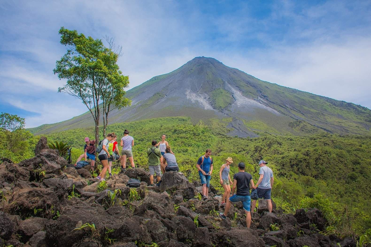 Excursionistas en un mirador rocoso frente a un imponente volcán rodeado de vegetación tropical.