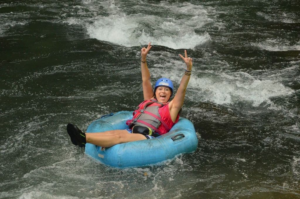 Mujer sonriente con casco y chaleco bajando por un río en un flotador azul inflable.